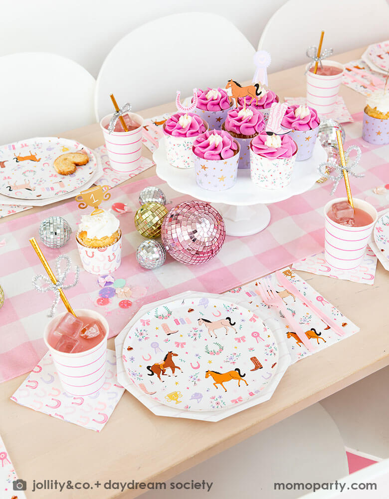 A girly cowgirl rodeo pony themed party table featuring Momopartys's cowgirl pony themed party plates, napkins, horseshoe napkins, pink striped party cups and a pink gingham table runner. Around the tableware there are disco ball decorations and a cake stand with cupcakes with pink icing, making this an adorable inspiration for girl's First Rodeo first birthday celebration or an adorable cowgirl theme party for pony-loving girls!