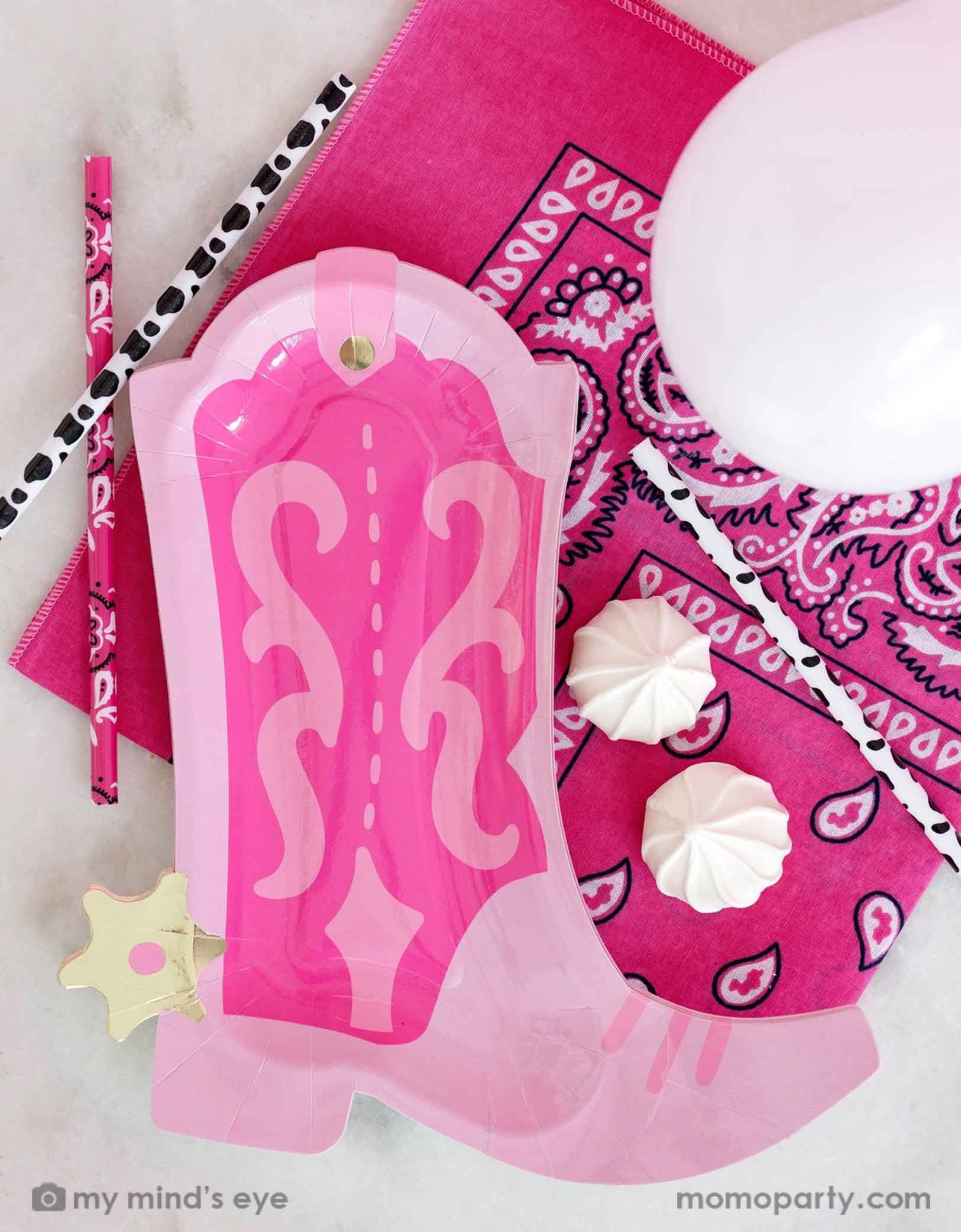 A overhead shot of a Western cowgirl themed party tablescape featuring Momopartys's cowgirl themed party supplies including the 10" x 11" pink cowgirl boot shaped plate, cow print paper straws, a pink bandana and some light pink balloons. Perfect for girl's firs rodeo first birthday celebration or a cowgirl disco themed bash.
