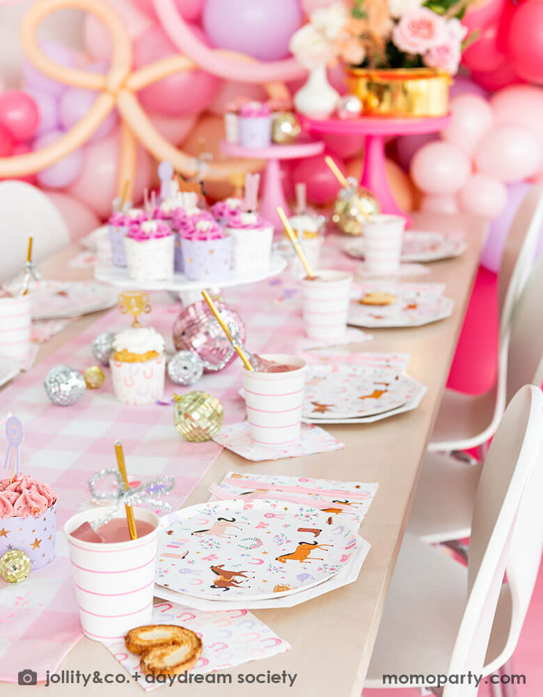 A close up of a girly cowgirl rodeo pony themed party table featuring Momopartys's cowgirl pony themed party plates, napkins, horseshoe napkins, pink striped party cups and a pink gingham table runner. Around the tableware there are disco ball decorations and a cake stand with cupcakes with pink icing. Behind the table there is a balloon wall with pink balloons, making this an adorable inspiration for girl's First Rodeo first birthday celebration or an adorable cowgirl theme party for pony-loving girls!