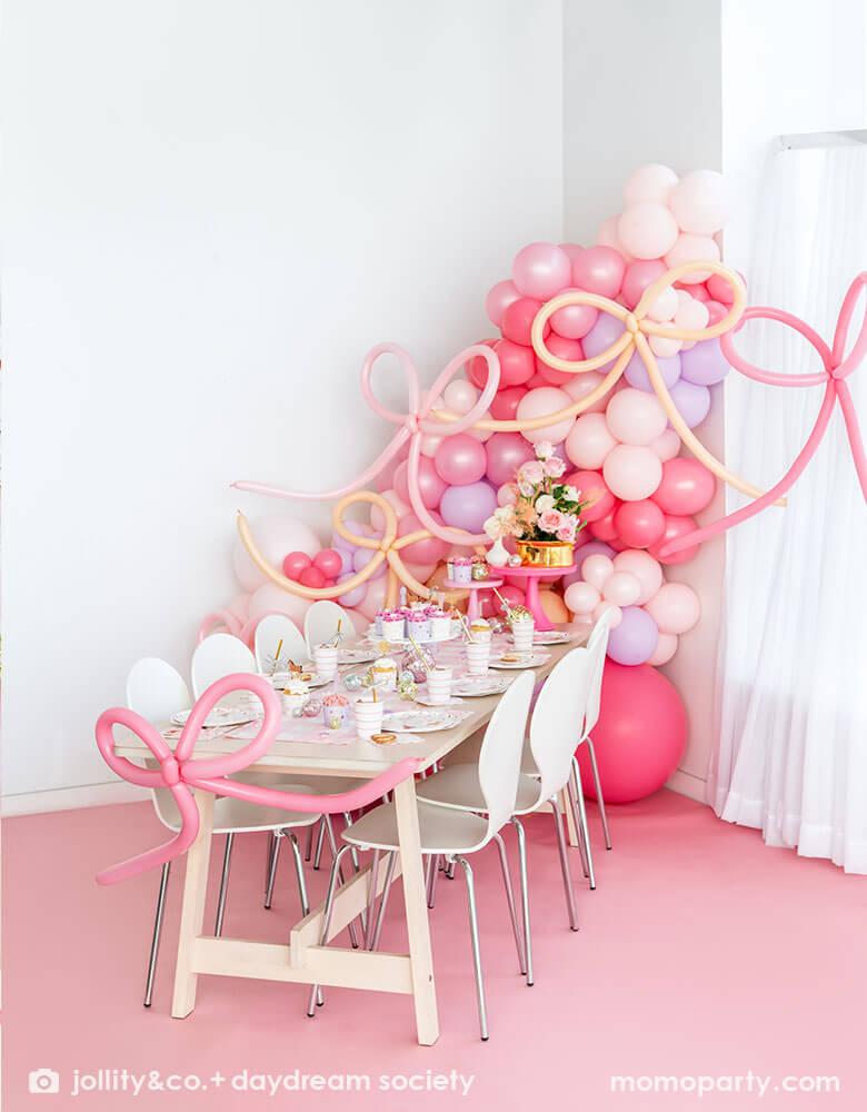 A beautiful girly pony themed birthday party set up featuring Momopartys's cowgirl pony themed party supplies and decorations on the party table. Around the tableware there are disco ball decorations, cupcakes with pink icing and pink floral bouquet. Behind the table there is a balloon wall with pink balloons adorned with pink bow shaped balloons, making this an adorable inspiration for girl's First Rodeo first birthday celebration or an adorable cowgirl theme party for pony-loving girls!