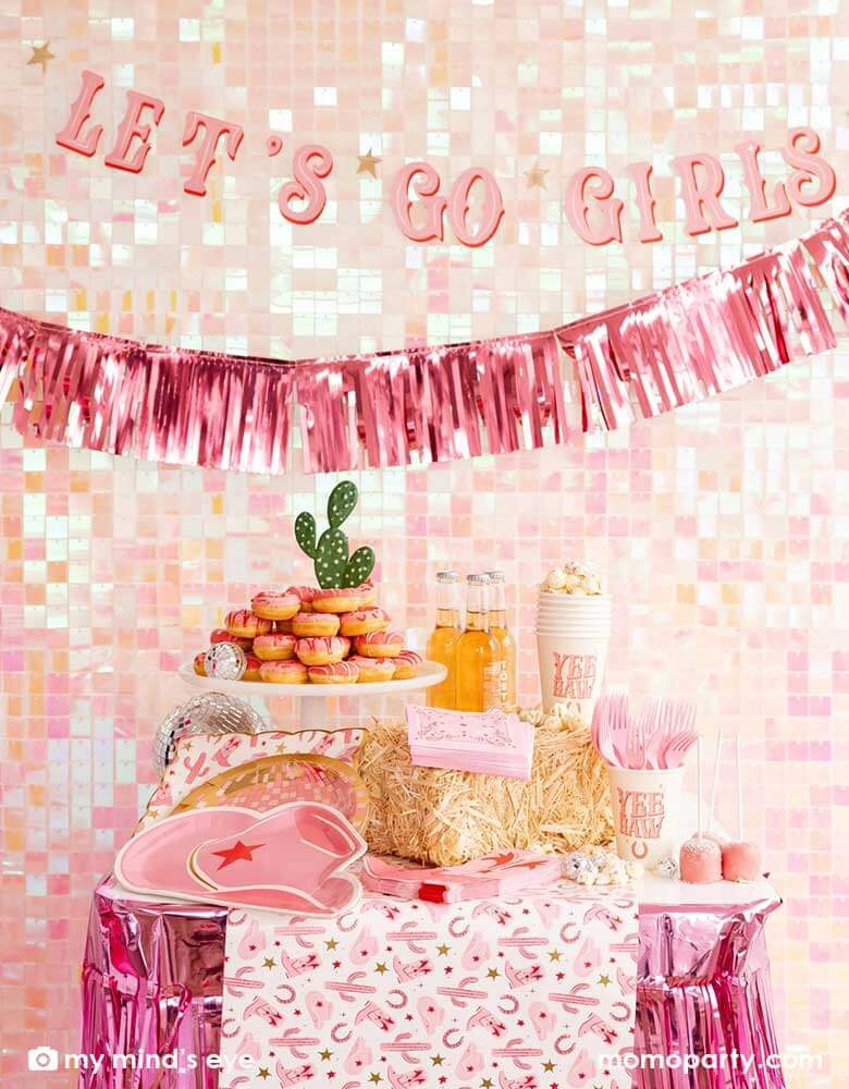 A cowgirl Western themed party table features pink cowgirl themed party supplies from Momopartys including pink cowgirl boot shaped napkins, cowgirl hat shaped plates, pink disco ball shaped plates, pink bandana small napkins, yeehaw party cups and cowgirl pattern paper plates and table runner by My Mind's Eye. In the back there's an iridescent backdrop adorned with Let's Go Girls pink foil fringe banners, along with a pink donut tower cake, making this a great inspo for a disco cowgirl party.