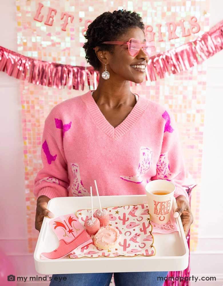 A young African American lady in a pink sweater with cowgirl boot pattern on it with disco ball ear rings and pink heart shaped sunglasses on her holding a tray with cowgirl party supplies from Momopartys including cowgirl pattern plates, pink cowgirl boot shaped napkins, yeehaw party cups and some pink treats. In her back there's an iridescent curtain wall adorned with My Mind's Eye's Let's Go Girls party banner with pink foil fringe banner, making this a great inspo for a disco cowgirl bachelorette party.