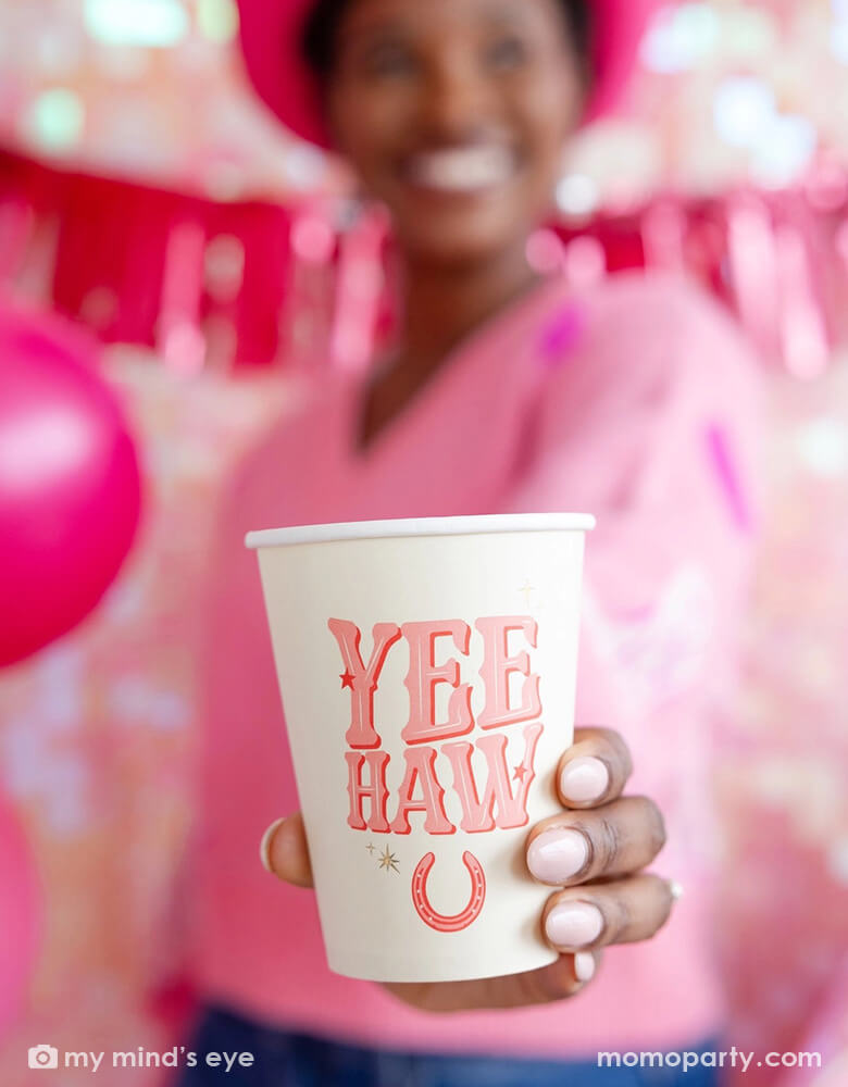 A young lady standing in front of a wall decorated with pink party banners and balloons, he's wearing a pink sweater with a pink cowgirl hat holding the 12 oz YEEHAW cowgirl party cup from Momopartys, enjoying herself in this disco cowgirl themed party.