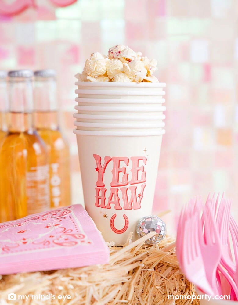 A cowgirl rodeo Western themed party table features pink cowgirl themed party supplies from Momopartys including 12oz pink YEEHAW party cups with popcorn in them, pink bandana small napkins and some soda bottles on a stack of hays, making this a perfect inspo for kid's cowgirl themed birthday party or a disco cowgirl bachelorette party celebration!