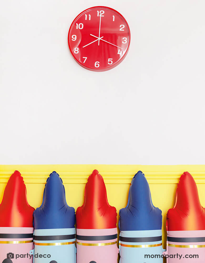 A line up of Momopartys's blue crayon shaped foil balloon and red crayon shaped foil balloon by Party Deco against a yellow wall. On the wall there's a red school clock at 8 oclock, creating a first day of school scene for kid's back to school season.