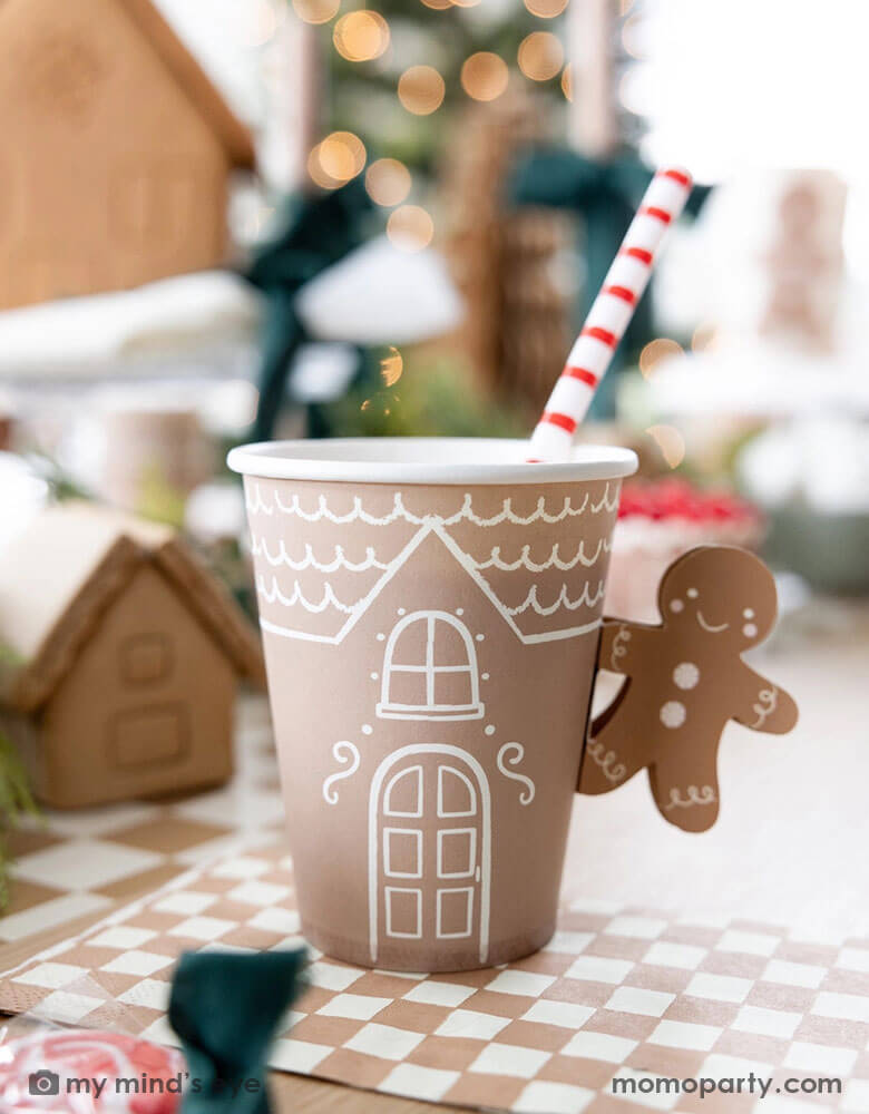 A festive gingerbread themed Holiday party table set up featuring gingerbread themed party tableware by Momopartys including the 12 oz Gingerbread Handled Paper Cup with a festive gingerbread man handle, the mocha checkered napkins and paper table runner. With gingerbread houses and Christmas trees in the back this creates a cozy yet festive vibe for a whimsical Holiday party for kids.