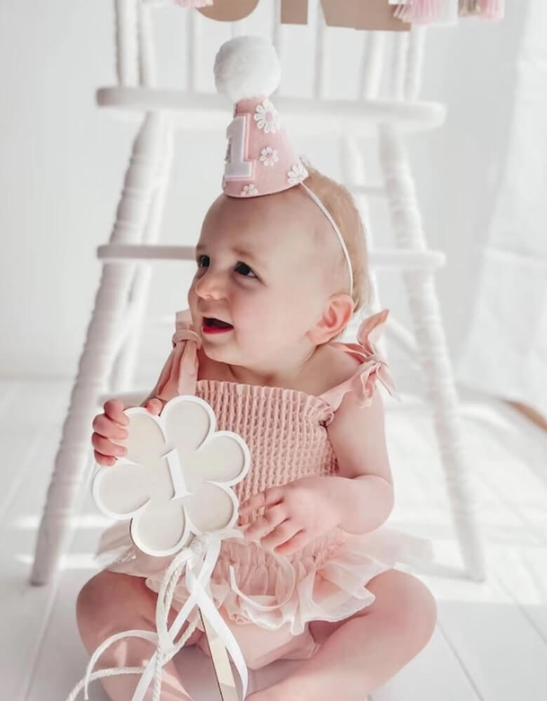 A little baby girl in adorable pink onesie wearing Momopartys's Daisy Applique felt birthday hat on her head. In her hands, she is holding a flower shaped pennant with number 1 on it. She's sitting in front of a vintage inspired white highchair celebrating her first birthday.