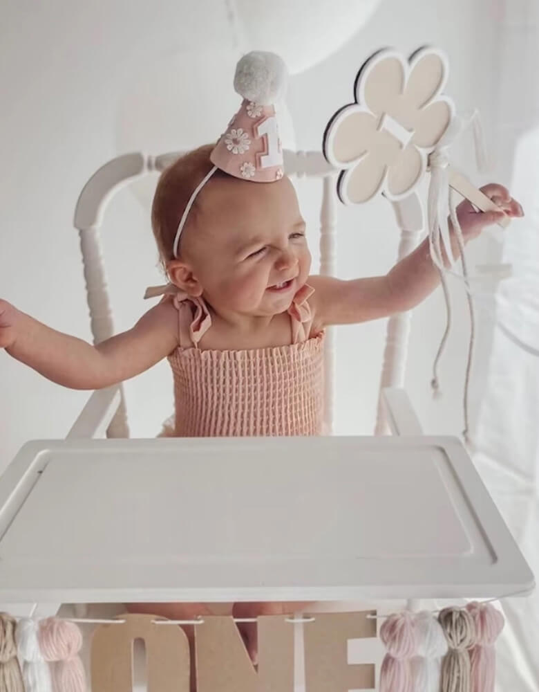 A happy baby girl in an adorable pink onesie wearing Momopartys's Daisy Applique felt birthday hat on her head. In one of her hands, she is holding a flower shaped wooden wand with number 1 on it. She's happily waving the want sitting on a white vintage inspired highchair which is adorn with ONE wool tinsel highchair garland  celebrating her first birthday.