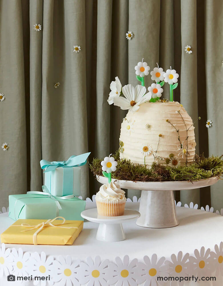 A wild daisy themed birthday party table featuring a buttercream cake decorated with real daisies on the side and topped with Momopartys's daisy candles from Meri Meri. There's also a daisy paper decoration wrap around the table which has some wrapped gifts on it.  In the back is a curtain decorated with real daisies flowers, making this an adorable inspo for girl's daisy or garden themed birthday party.
