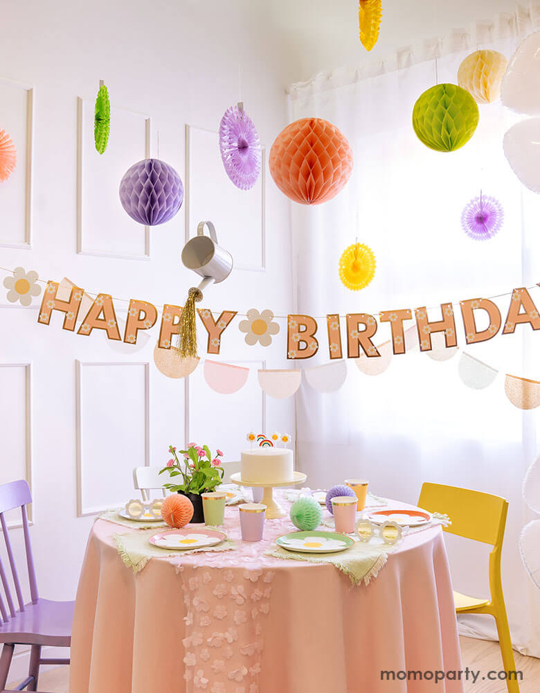 A bright and clean dining room decorated with pastel colored honeycombs and party fans hung from the ceiling. Momopartys's daisy Happy Birthday banner is hung underneath. On the table there are a various daisy themed tableware including plates, cups and birthday candles form Momopartys. Making this a dreamy, whimsical party set up for girl's birthday.