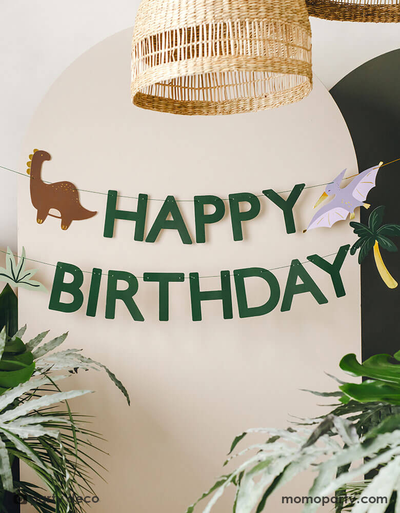 A kid's dinosaur themed party backdrop wall decorated with Momopartys's dinosaur happy birthday banner by Party Deco. This green happy birthday banner featuring dinosaur and palm tree pennants is perfect to set a scene for kid's dinosaur, Jurassic themed birthday party.