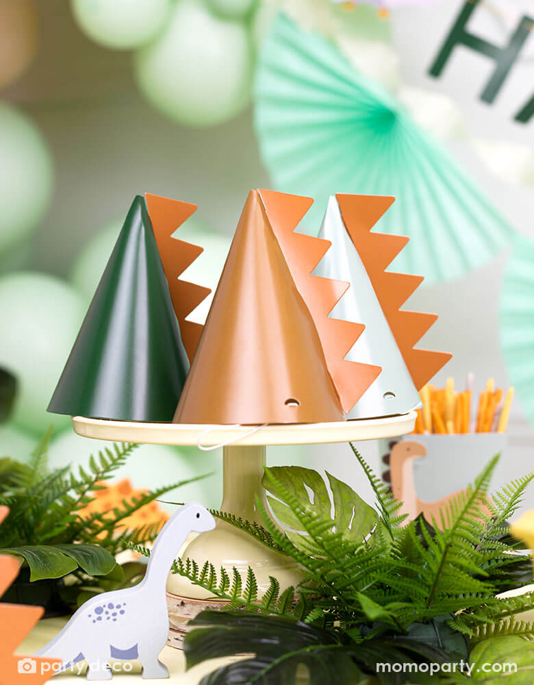 A festive kid's dinosaur themed party table features Momopartys's 5.5" Dinosaur Party Hats by Party Deco. Comes in a set of 6 party hats in 3 different colors of green, mint, and brown, these dinosaur party hats are with a dinosaur spike on them. The table is decorated with jungle themed plants with mint, brown and green colored decorations on the wall including some party balloons and a festive bunting garland.