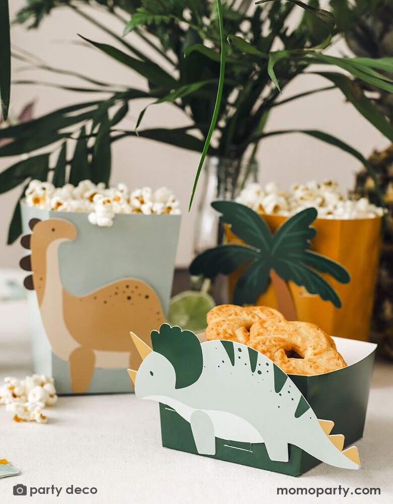 A kid's dinosaur party table features Momopartys's dinosaur snack boxes filled with cookies and popcorn, around the table are some tropical plants, creating a Jurassic pre-historic vibe for this dinosaur adventure.