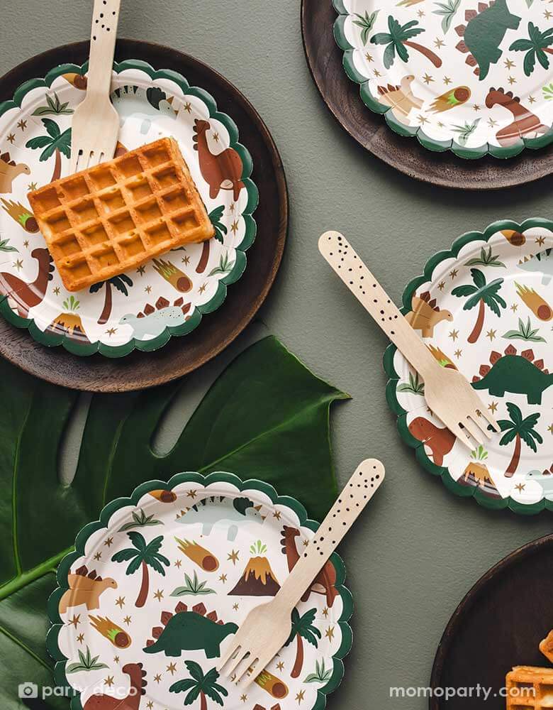 A dinosaur party table featuring Momopartys's 7" scalloped dinosaur side plates  in olive green background and palm leaves  around. With waffle snacks and wooden knives, these adorable dinosaur party plates are perfect for kid's dinosaur themed birthday celebrations.