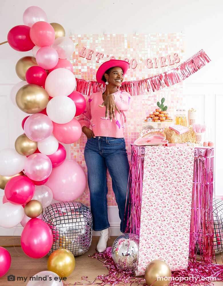 A young African American lady in the Western inspired outfits and a pink cowgirl hat in a disco cowgirl bachelorette party decorated with cowgirl themed party supplies, decorations and balloons from Momopartys. She's showing her engagement ring next to a table adorned with pink fringe and cowgirl party supplies including plates, napkins and a paper table runner. There's a donut cake, beverages and various treats on the table. Behind them is a iridescent wall with "Let's Go Girls" banner from Momopartys.