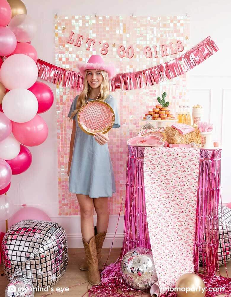 A young Caucasian lady in the Western inspired outfits and a pink cowgirl hat in a disco cowgirl bachelorette party decorated with cowgirl themed party supplies, decorations and balloons from Momopartys. She's holding the pink disco ball shaped plate by a table decorated with cowgirl party supplies including plates, napkins and a paper table runner. There's a donut cake, beverages and various treats on the table. Behind them is a iridescent wall adorned with "Let's Go Girls" pink foil fringe party banner.
