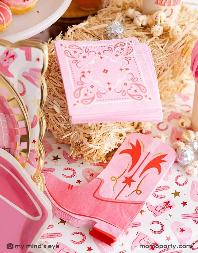 A cowgirl rodeo Western themed party table features pink cowgirl themed party supplies from Momopartys by My Mind's Eye including pink cowgirl boot shaped napkins, pink bandana small napkins, cowgirl hat shaped plates, yeehaw party cups and cowgirl pattern paper table runner, along with hay stacks and small disco ball decorations, this makes a perfect inspo for kid's cowgirl themed birthday party or a bachelorette party celebration!