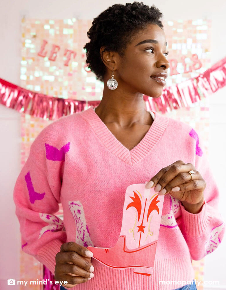A young African American lady in a pink sweater with cowgirl boot pattern on it with disco ball ear rings on her holding Momopartys's pink cowgirl boot napkins. In her back there's an iridescent curtain wall adorned with My Mind's Eye's Let's Go Girls party banner with pink foil fringe banner, making this a perfect inspo for disco cowgirl bachelorette party celebration.