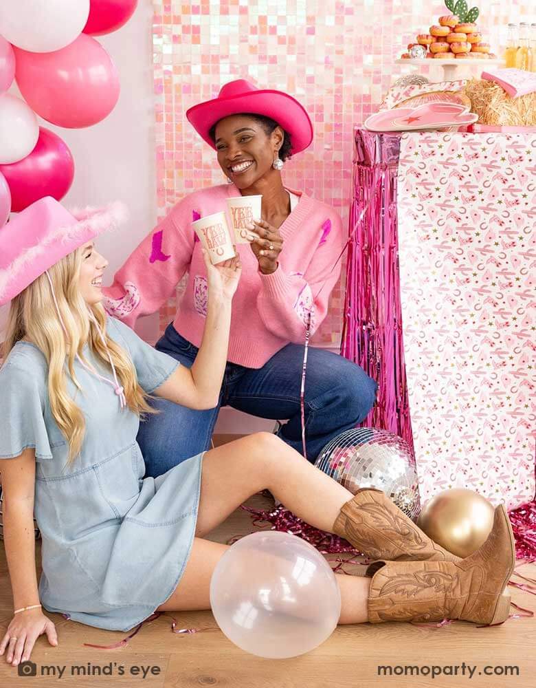 Two ladies in their Western rodeo cowgirl inspired outfit in a disco cowgirl themed bachelorette party decorated with cowgirl themed party supplies, decorations and balloons from Momopartys. They're raising their 12oz YEEHAW party cups by My Mind's Eye enjoying themselves in this festive celebration with donut cakes, beverages and various treats on the table decorated with cowgirl pattern table runner and pink foil fringe next to them. 