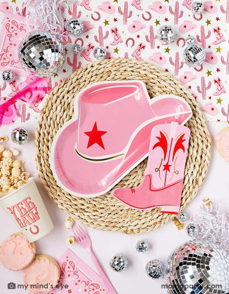 A cowgirl rodeo Western themed party table features pink cowgirl themed party supplies from Momopartys including pink cowgirl boot shaped napkins, cowgirl hat shaped plates, pink bandana small napkins, yeehaw party cups and cowgirl pattern paper table runner, along with festive disco ball decorations, heart shaped sunglasses and treats, this makes a perfect inspo for kid's cowgirl themed birthday party or a bachelorette party celebration!