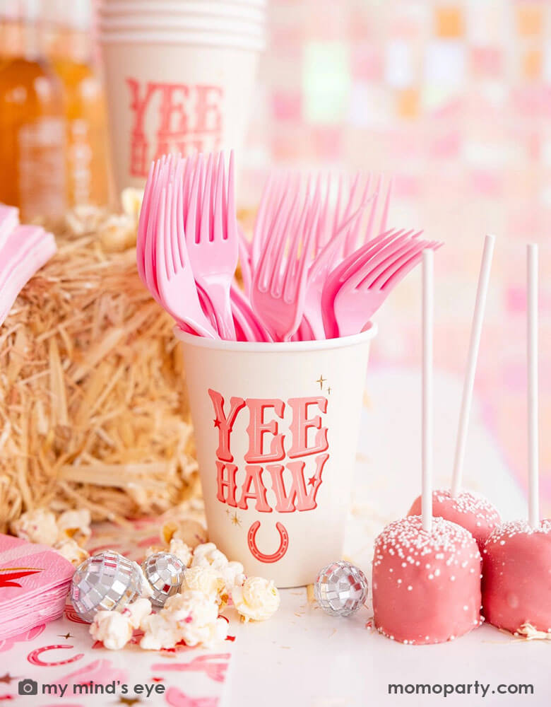 A cowgirl rodeo Western themed party table features pink cowgirl themed party supplies from Momopartys including 12oz pink YEEHAW party cups with pink utensils, cowgirl boot shaped napkins on the cowgirl pattern paper table runner, along with hay stacks, some small disco ball decorations and pink cake pops and popcorn, making this a perfect inspo for kid's cowgirl themed birthday party or a disco cowgirl bachelorette party celebration!