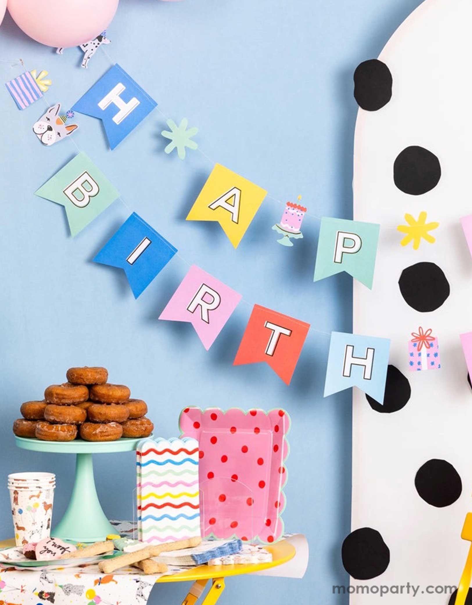 A corner of a festive and cheerful dog themed party featuring Momopartys's dog party tableware collection including dog themed party tablewares, the dog party Happy Birthday banner hung over a polka dotted party backdrop and party balloons. On the table there are some sugar cookies, treats and a donut tower,  ready for a fun and playful birthday celebration for a pet lover!