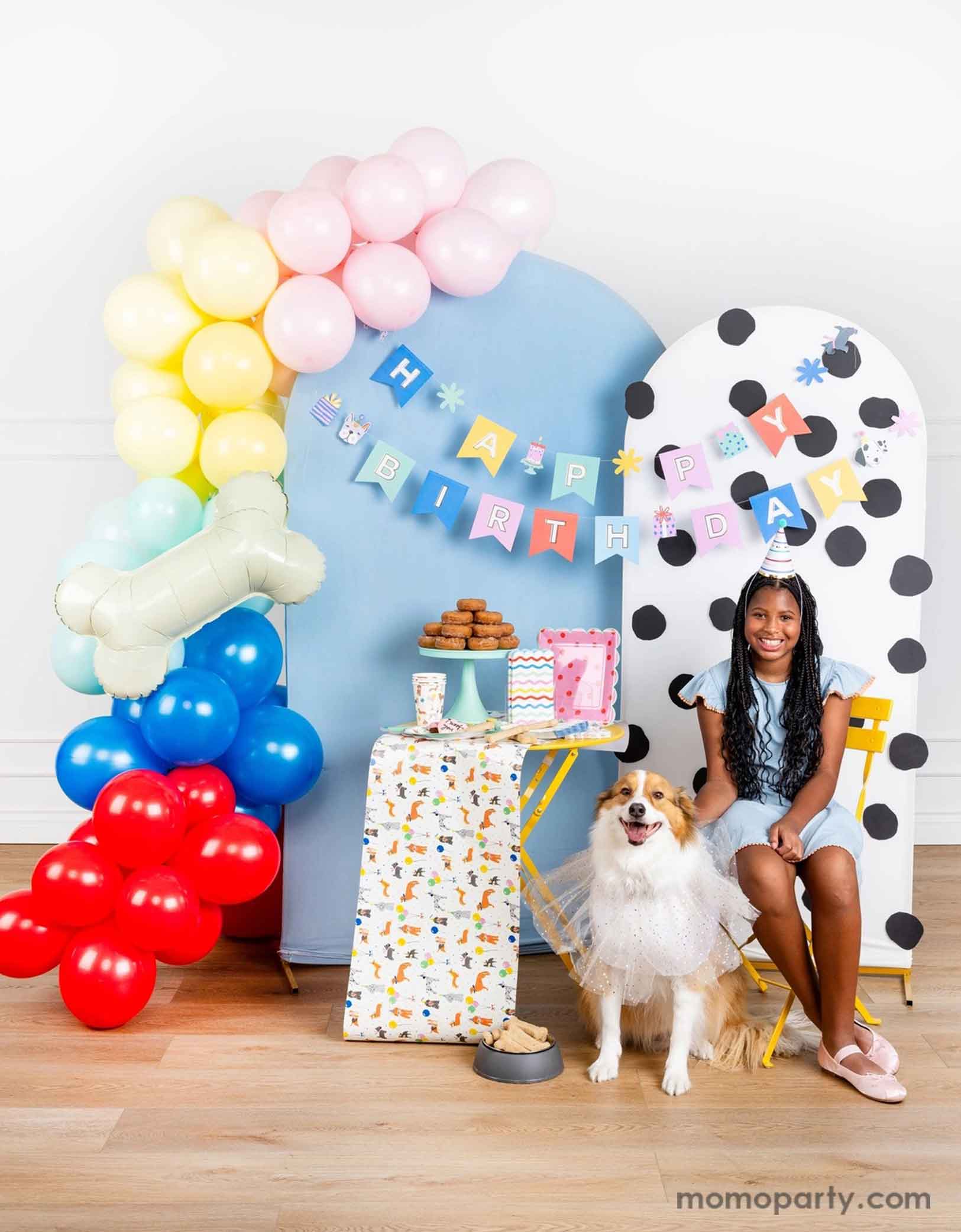 A dog themed party decoration set up featuring Momopartys's dog themed birthday supplies by My Mind's Eye. Including dog Happy Birthday party banner hung over a polka dotted and a pastel blue party backdrop with a colorful party balloon garland and a bone shaped foil balloon. On the table there are some dog themed party tablewares sugar cookies, treats and a donut tower. A girl in a birthday hat is sitting on the side with her dog happily celebrating her birthday.