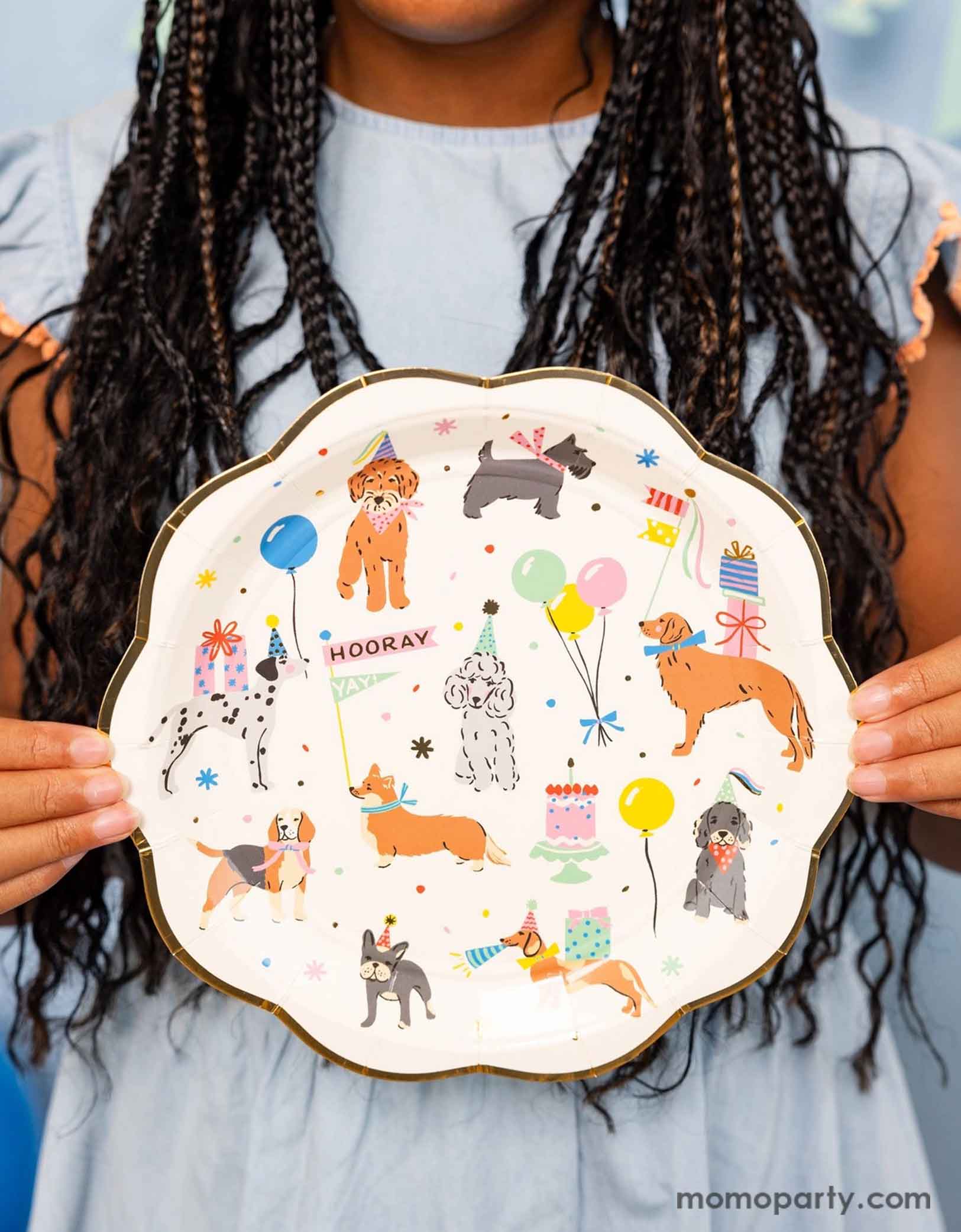 A girl in her pastel blue dress holding Momopartys's  9" Dog Party Plates by My Mind's Eye. Featuring adorable illustrations of different breeds of dogs with party hats, balloons and birthday cakes, these playful dog paper plates are perfect for any pet themed celebration.