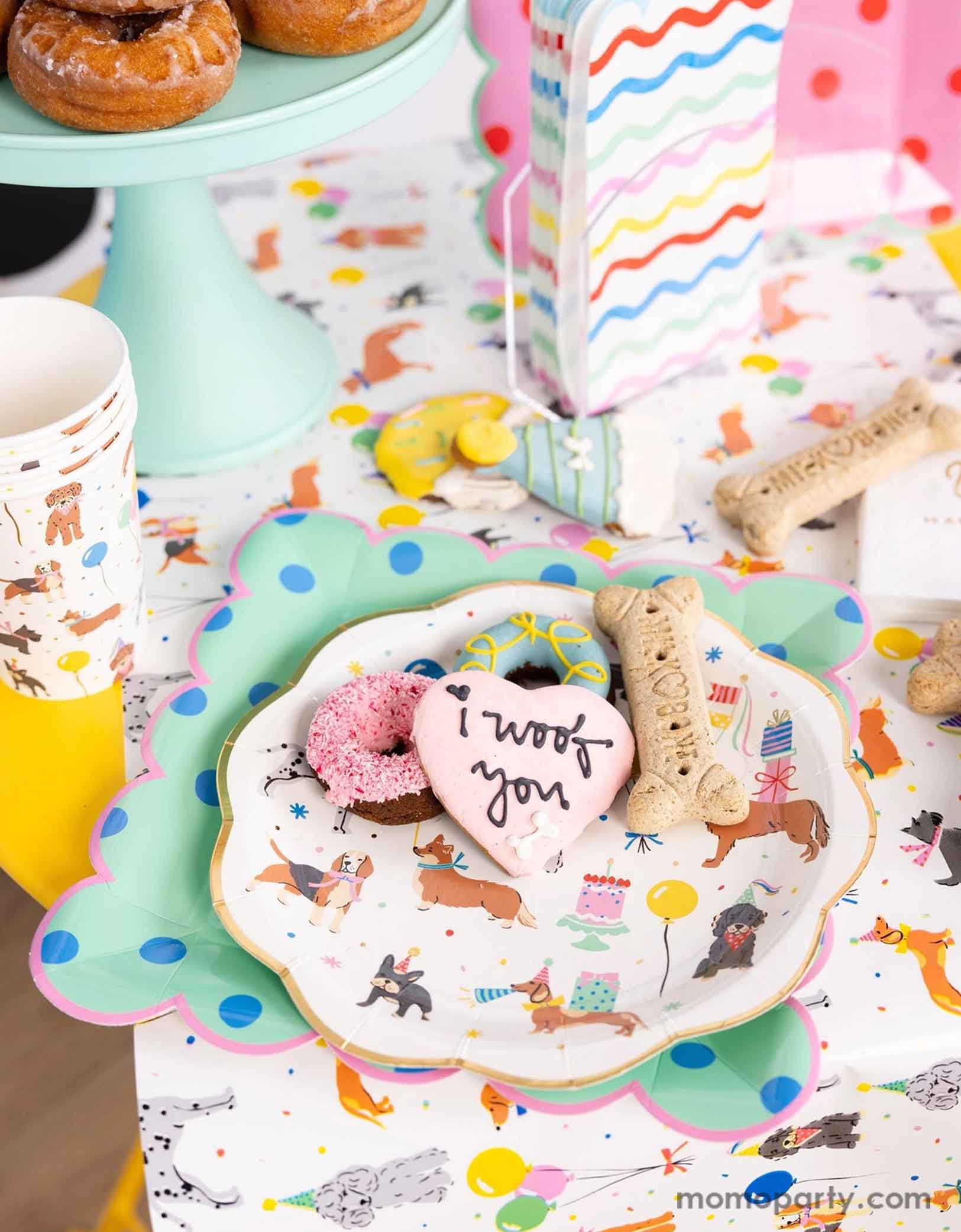 A festive and cheerful dog themed party table featuring Momopartys's dog party tableware collection including dog paper plates, party cups, napkins and table runner featuring adorable illustrations of different breeds of dogs with party hats, balloons and birthday cakes. With mint polka dot square plates and birthday festive sugar coated cookies that say "I Woof You" and donuts. This table is ready for a fun and playful birthday celebration for a pet lover!