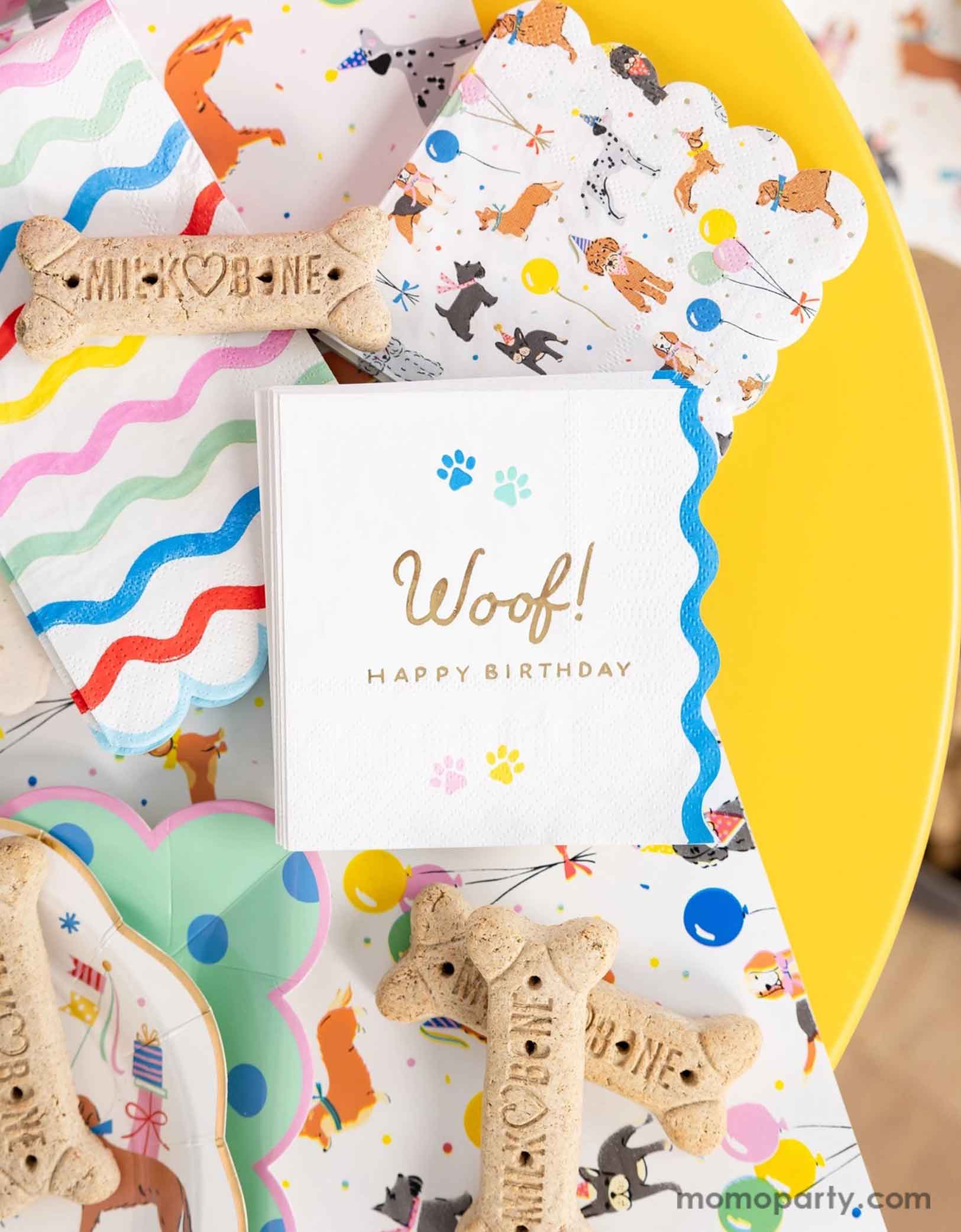 A festive and cheerful dog themed party table featuring Momopartys's dog party tableware collection including dog paper plates, woof! Happy Birthday napkins and table runner featuring adorable illustrations of different breeds of dogs with party hats, balloons and birthday cakes. With mint polka dot square plates and rainbow ric rac guest napkins, along with dog bone shaped cookies, this table is ready for a fun and playful birthday celebration for a pet lover!