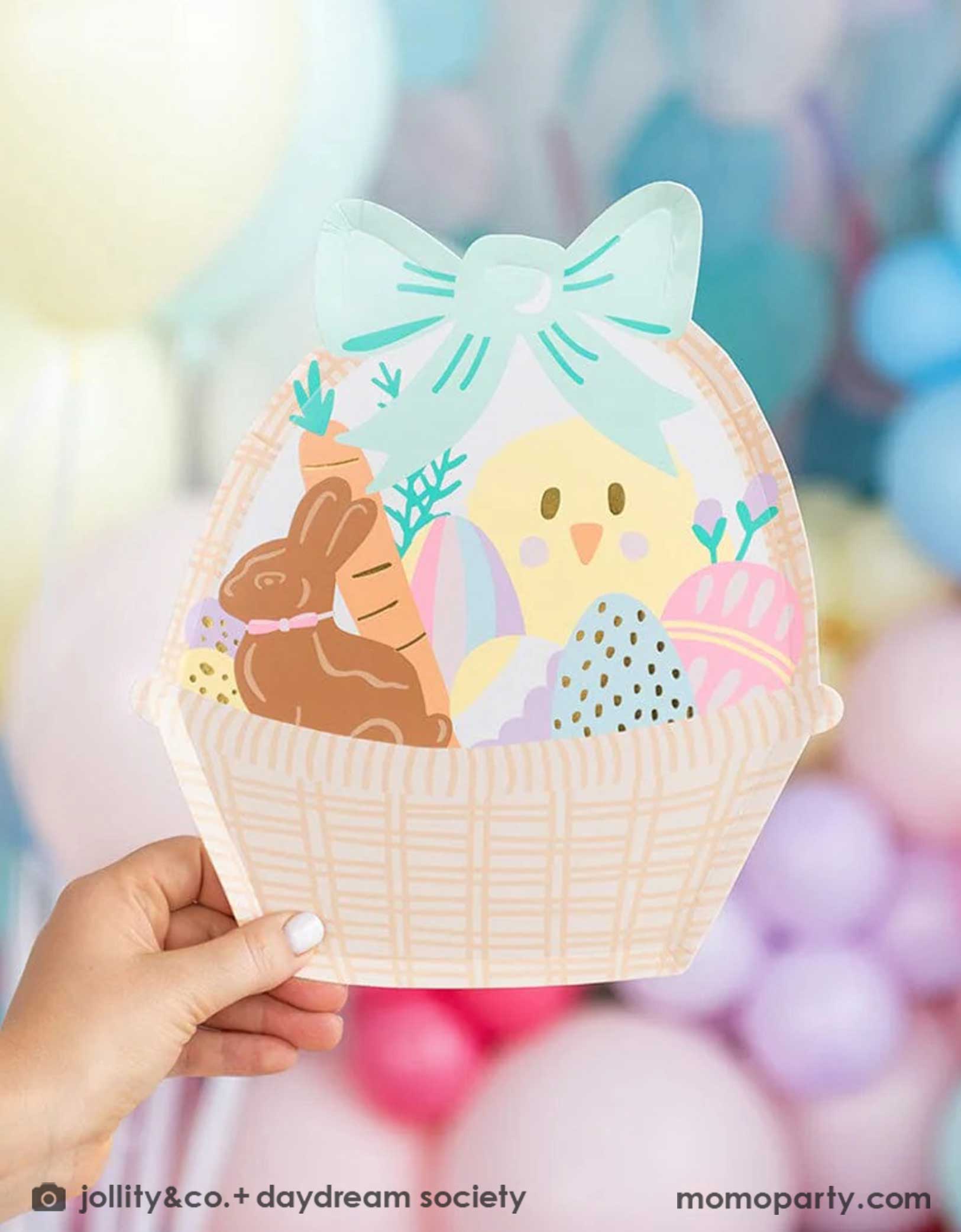 A little kid's hand holding Momopartys's Easter Fun basket shaped plate by Daydream Society. In the back there's a wall of balloons in pastel spring inspired colors, making this an adorable Easter party decoration.