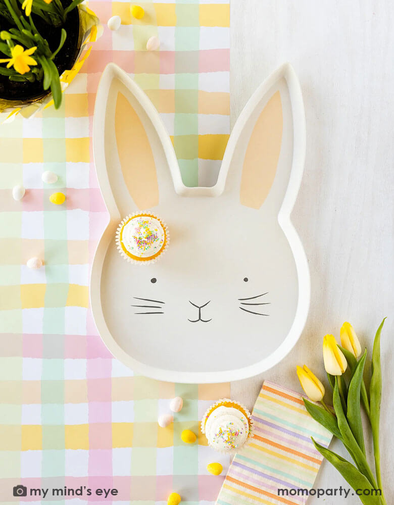 An Easter bunny shaped tray with sprinkled cupcakes on Momopartys's Momopartys's 16 x 120 inches Spring Checks Paper Table Runner by My Mind's Eye. Around it are yellow tulips and pastel striped guest napkins and spring colored jelly beans - bringing all the spring vibes to this Easter tablescape.
