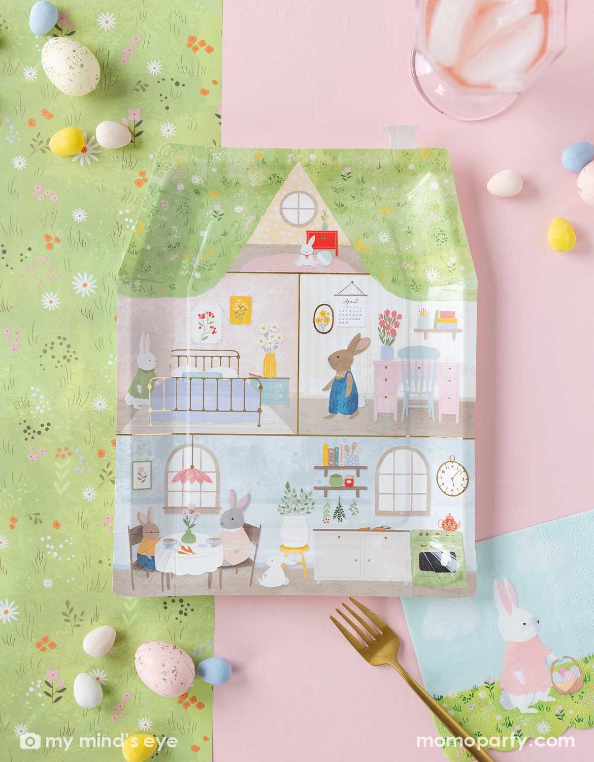 An overhead shot of an Easter-themed tablescape featuring Momopartys's bunny bunny house shaped plate and the matching napkin on a cottage meadow table runner with easter egg shaped candies scattered around.