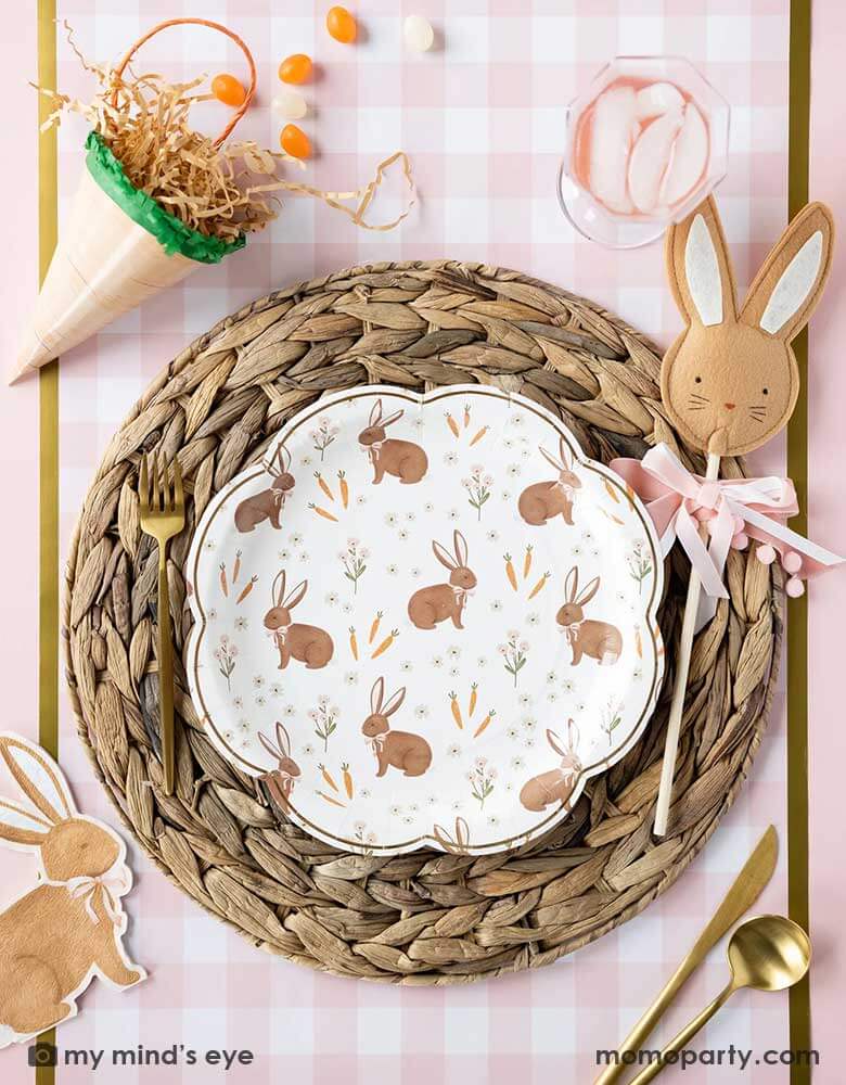 A beautiful Easter tablescape features Momopartys's scatter rabbit scallop paper plate by My Mind's Eye in the middle, laid on top of a straw woven placemat which is on the pink gingham checks paper table runner, around the plate are carrot shaped treat boxes, brown rabbit shaped napkin and Easter rabbit wand, makes this an adorable kid's friendly Easter tableset this spring.