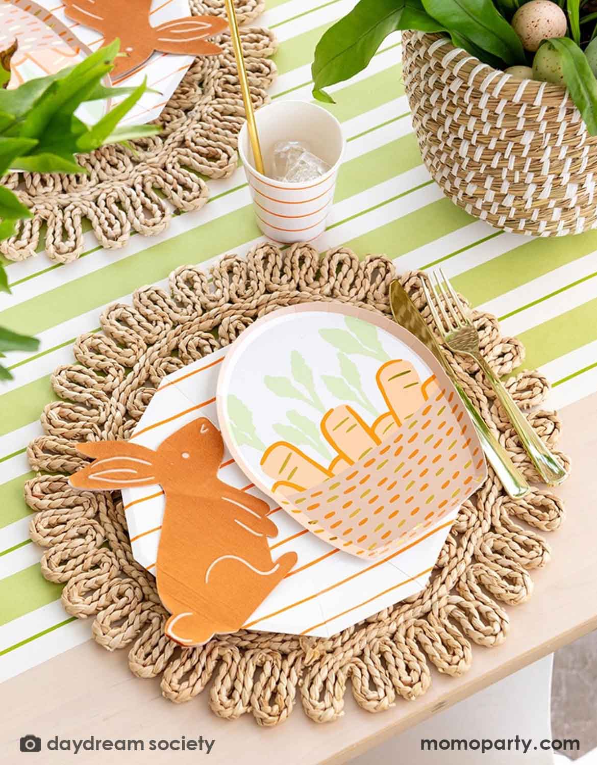 An Easter bunny themed table setting with Momopartys's bunny themed tablewares including carrot basket plates, bunny shaped napkins and matching cups and utensils on a straw-weaved placemats over a green and white striped table runner.