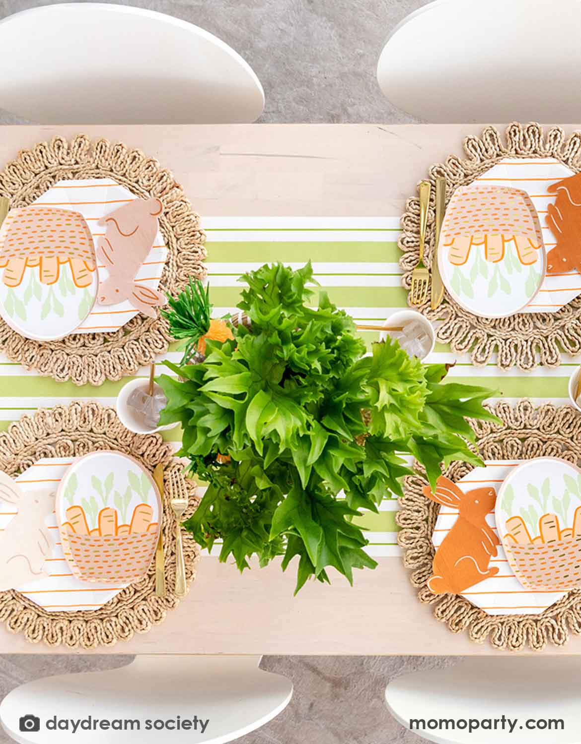 An overhead shot of an Easter bunny themed table setting with Momopartys's bunny themed tablewares including carrot basket plates, bunny shaped napkins and matching cups and utensils on a straw-weaved placemats over a green and white striped table runner. In the center there's a green and carrot floral arrangement as the centerpiece.