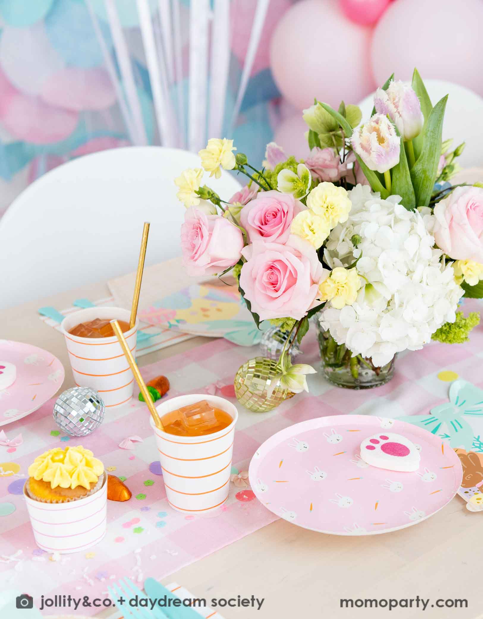 A beautiful Easter party table features Momopartys's Easter fun small plates along with striped party plates, cups, and napkins. In the middle of the table lays a pink gingham table runner which has a beautiful spring flower arrangement on it. In the back of the table there are some pastel balloons and paper decorations, making this a great look and inspo for a kid's friendly Easter party decoration.
