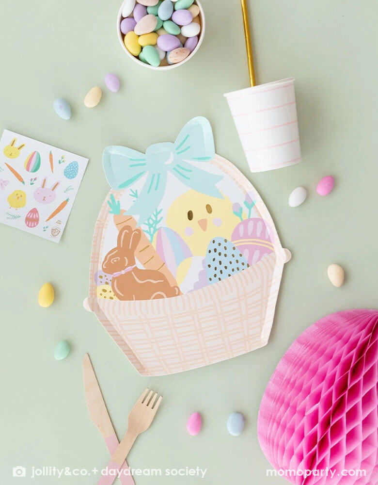 A pale mint table features Momopartys's 9.5 x 10.75 inches Easter basket shaped large plates by Daydream Society. Around it are some easter egg shaped candies, a light pink striped party cup, an Easter Fun sticker, pink wooden cutlery and sheet and a pink Easter egg tissue honeycomb.