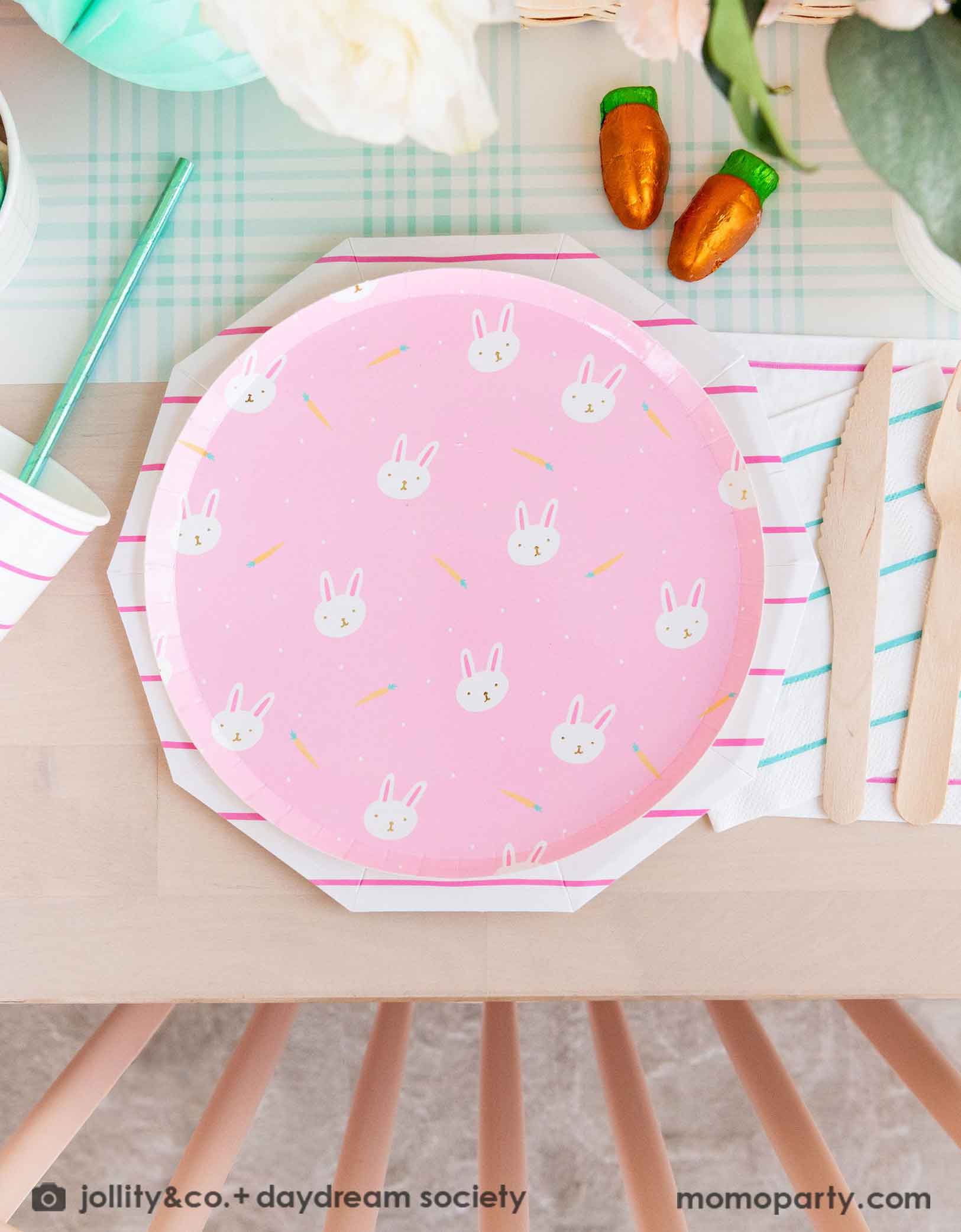 A beautiful Easter brunch table features Momopartys's pink Easter fun small plates along with striped party plates, cups, and napkins. In the middle of the table lays a blue gingham table runner which has baskets of spring flower arrangement and carrot shaped candies, making this an adorable inspo for a kid's friendly Easter party table setting.