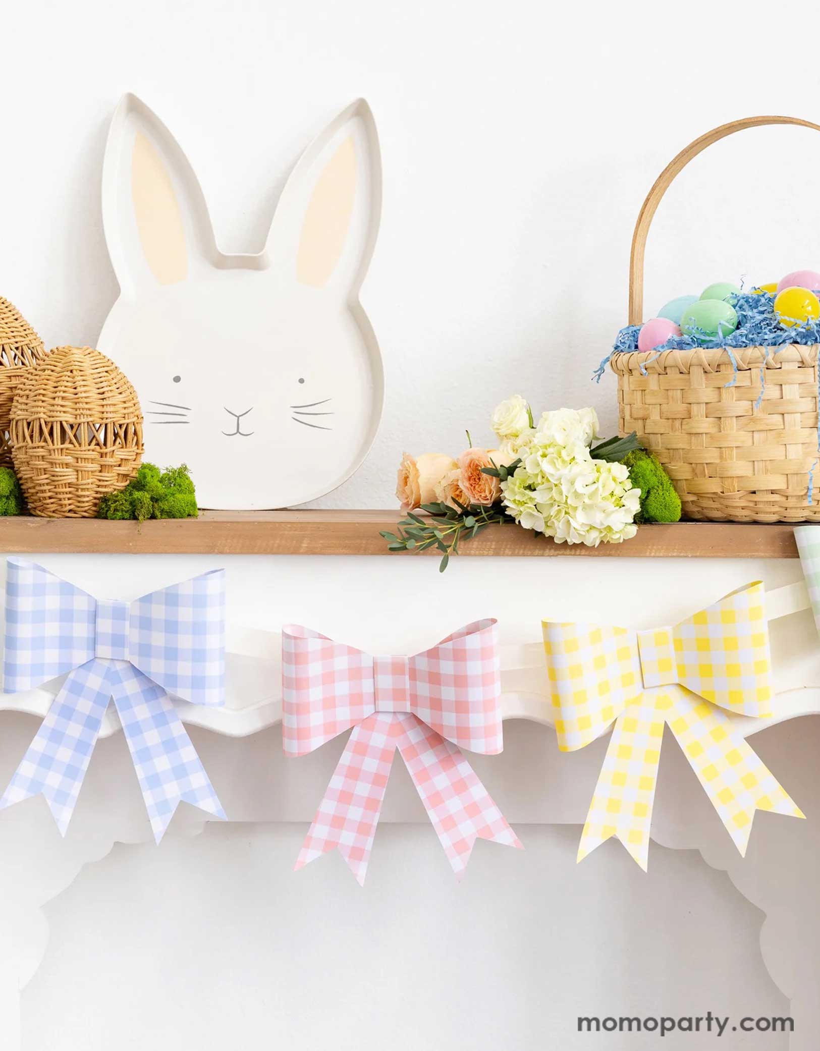 A mantel decorated with Easter decors including Momopartys's pastel gingham checkered bow party banner, Easter bunny shaped bamboo serving tray and some woven Easter egg decors, along with a basket filled with Easter eggs and spring floral bouquet, a simple yet adorable idea for spring home decor.