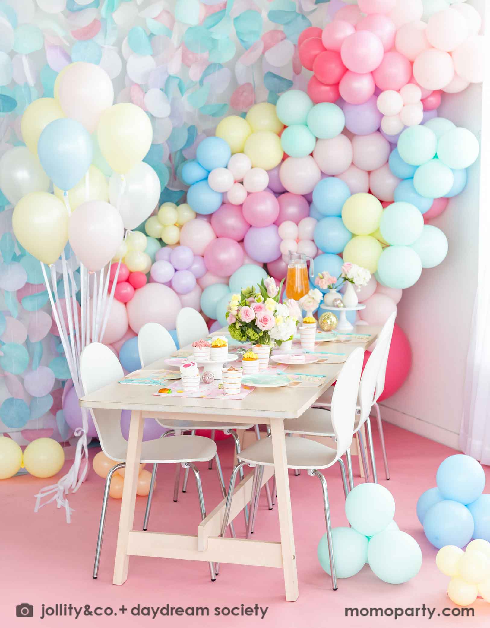 A festive Easter party table features Momopartys's Easter fun small plates along with striped party plates, cups, and napkins. In the back of the table there's a pastel balloon wall with pastel paper decorations, making this an adorable inspo for a Easter party decoration set up.