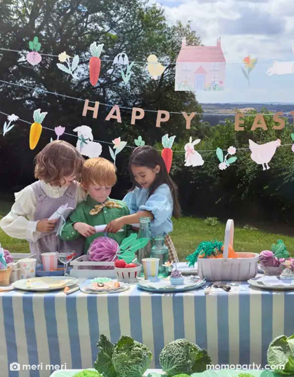 Children at an Easter-themed outdoor table setting with Meri Meri's 'Happy Easter' honeycomb banner and decorative elements hung above. On the table there are various Easter themed tableware and garden vegetable honeycombs including carrots, beets, and lettuces around, making it an adorable set up for kid's Easter celebration or garden themed birthday party in the spring time.  