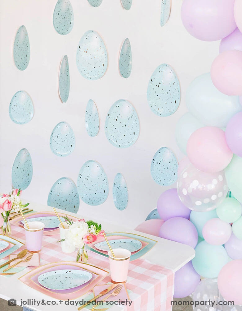A spring inspired Easter party table decoration featuring Momopartys's pastel themed tableware on the party table including pastel mint & pink plates and robin egg shaped plates, lilac party cups, pink gingham table runner and gold utensils. Next to the table there's a pastel balloon garland next to some robin egg party garland hung on the wall, making this a great Easter party inspo for springtime.