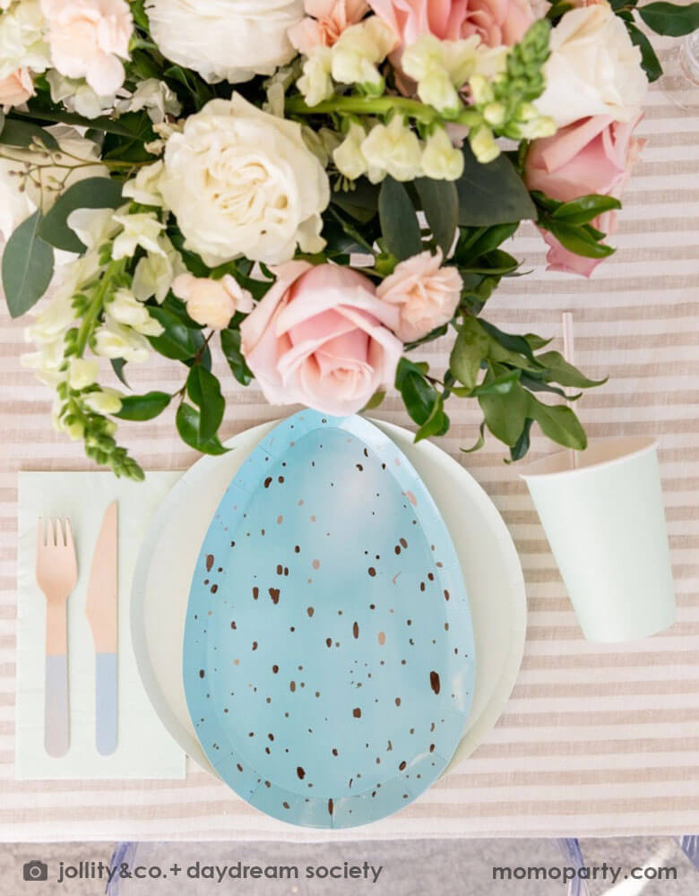 An closeup shot of Easter party table set up with Momopartys's Rockin' Robin Dinner Plates layered with pastel mint round plate, color matching paper cups, napkins, and pastel blue dipped wooden utensils, with a vase with flowers and on a light beige striped tablecloth for a modern chic Easter table placesetting.