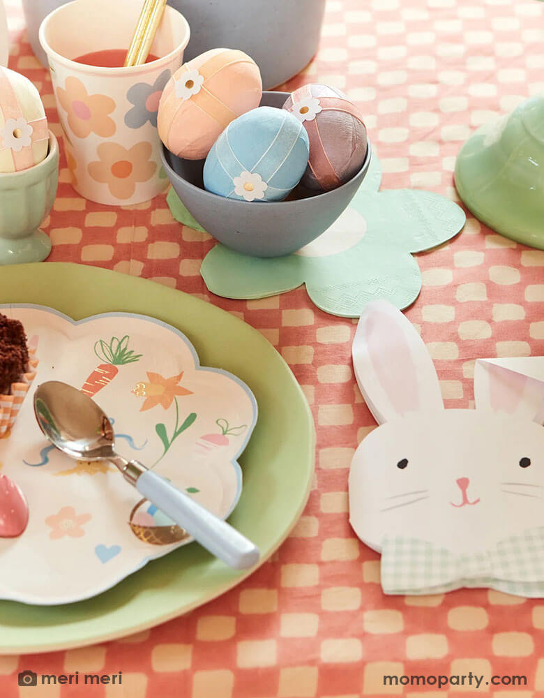 An Easter party table with Easter eggs adorned with Meri Meri's Easter themed tattoo stickers, on the table are Momopartys's Easter themed plates and napkins in soft pastel colors including bunny shaped napkins which are coordinated with the charming daisy shaped napkins, makes this an adorable tablescape idea for a spring gathering.