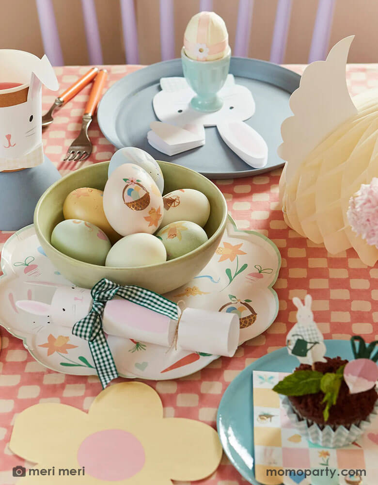 An Easter party table with Easter eggs adorned with Meri Meri's Easter themed tattoo stickers, on the table are Momopartys's Easter themed plates and napkins in soft pastel colors including bunny shaped napkins and party cups and bunny party crackers which are coordinated with the charming daisy shaped plates, makes this an adorable tablescape idea for a spring gathering.