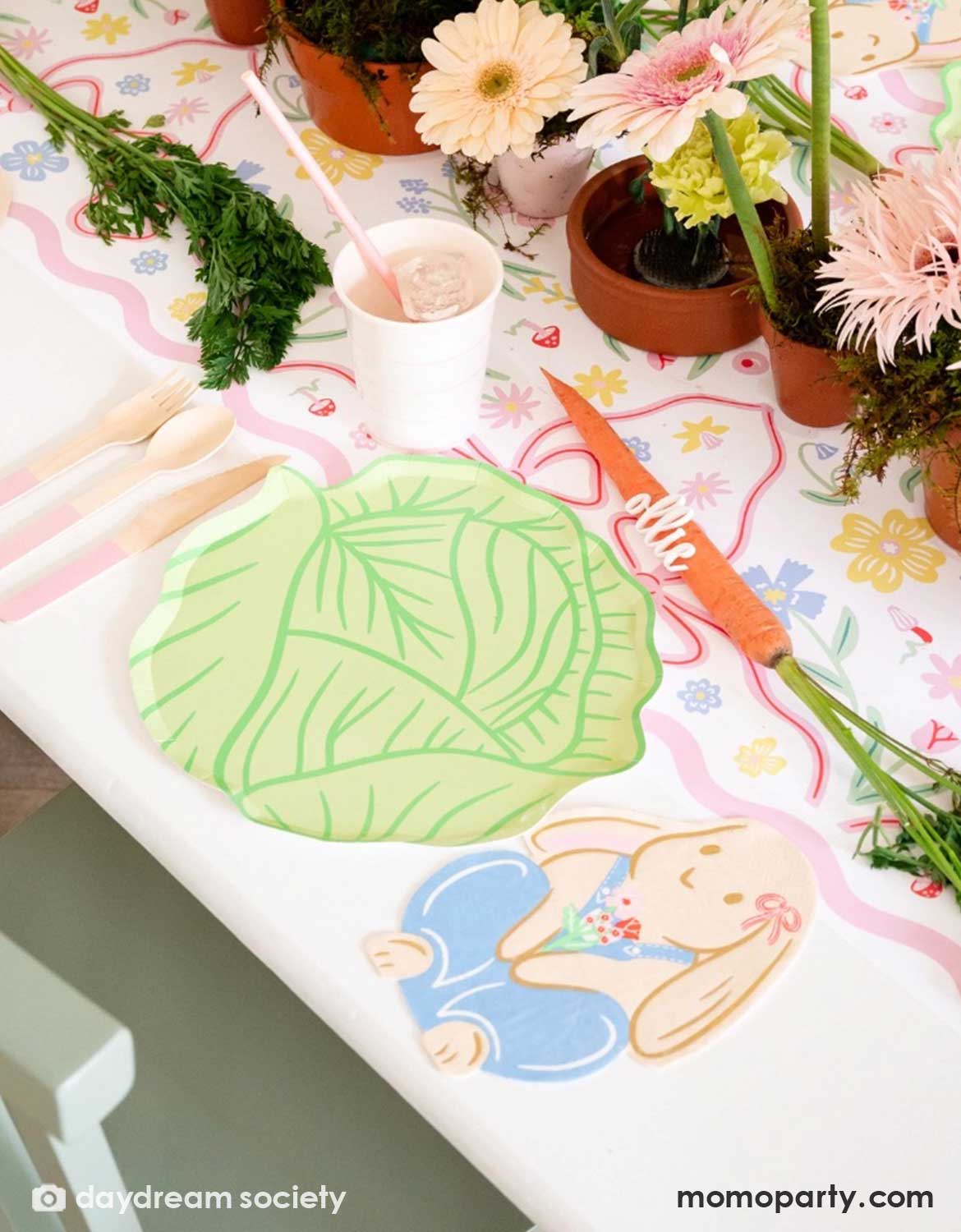 A spring inspired party table setting featuring colorful Easter themed tablewares from Momopartys including cabbage shaped plates, Easter bunny shaped napkins, matching party cups and utensils over a floral tablecloth with  potted plants in the middle as centerpiece.