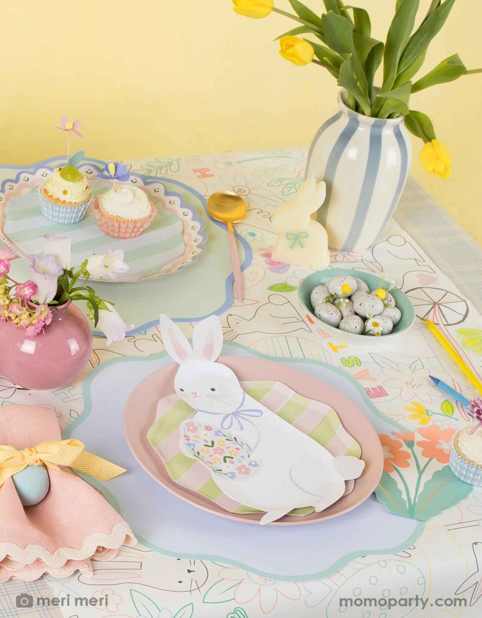 A kid's friendly Easter table setting featuring Momopartys's Easter party supplies by Meri Meri including the Easter bunny shaped plates, gingham checkered Easter cupcake kit, the Easter bunny coloring tablecloth, with pastel blue scalloped placemats, with spring tulip flowers and Easter egg shaped chocolate candies on the table, this makes a cute and simple placeset to celebrate Easter with your kids. 