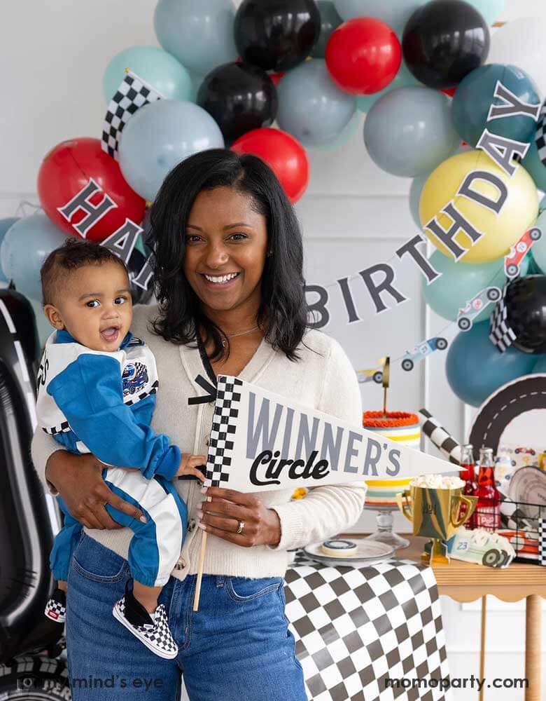 A mom holding his one year old boy in her arm standing in front of a race car themed party decoration set up which includes a colorful balloon garland in red, blue, gray black and yellow adorned with a happy birthday paper banner, on the table there are race car themed partyware including plates, napkins, checkered table runner and race car flags. The mom is holding a felt party pennant written "Winner's Circle" from Momopartys.