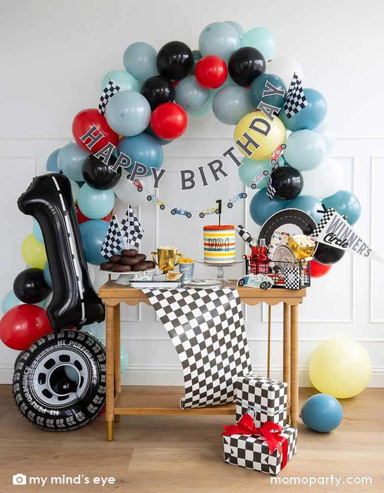 A "Fast One" boy's race car themed first birthday party decoration featuring festive race car themed party supplies from Momopartys including Miles per Hour party plates, cups, napkins, placemats, trophy treat cups, and a checkered table runner. On the party table there are a cake and donuts topped with checkered flags. Above the table is an organic balloon garland adorned with race car happy birthday banners. Along with a tire shaped foil balloon a number one letter balloon with decorated with road tape.
